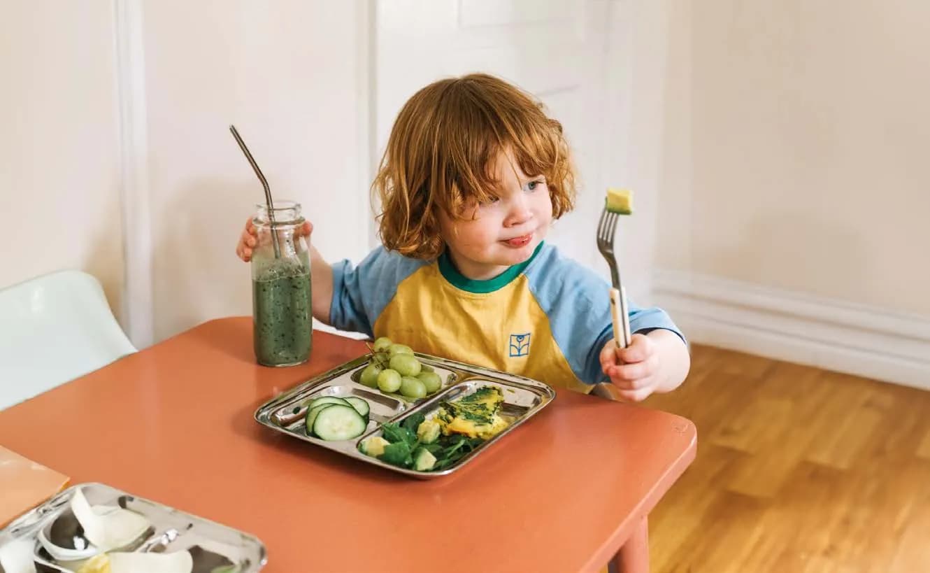 Un jeune enfant attablé, fourchette en main, devant un plateau de légumes et de raisins.